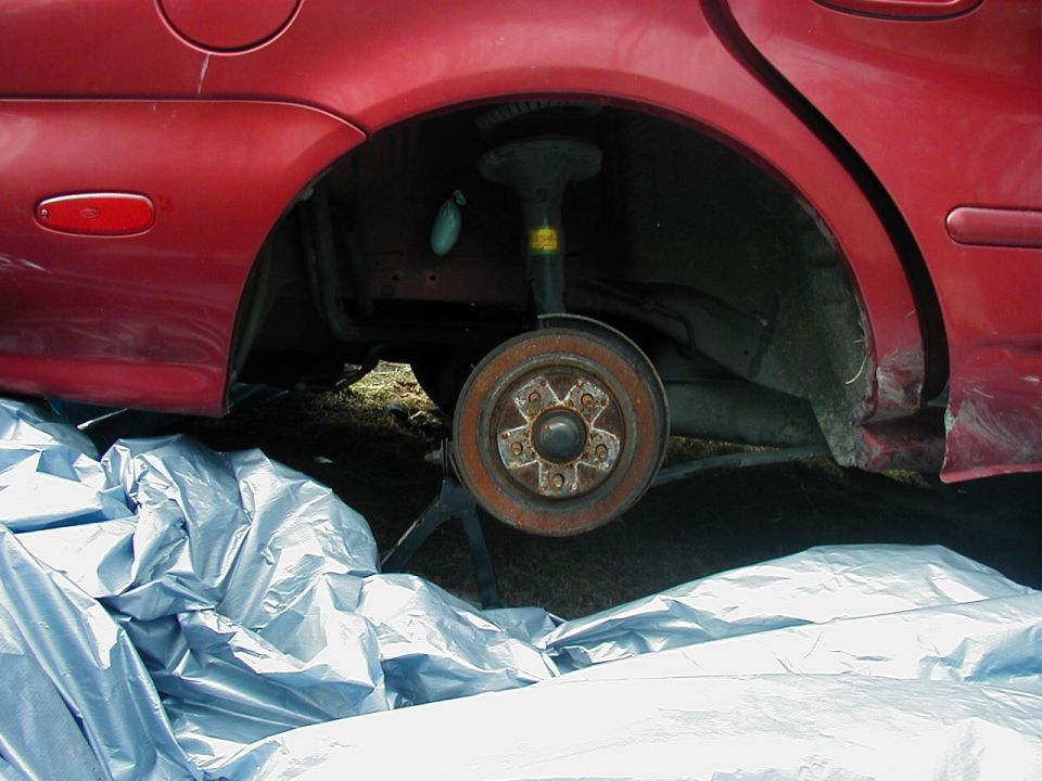 Red car's rear wheel brake assembly removed, supported by a jack stand. Blue tarp covers the ground