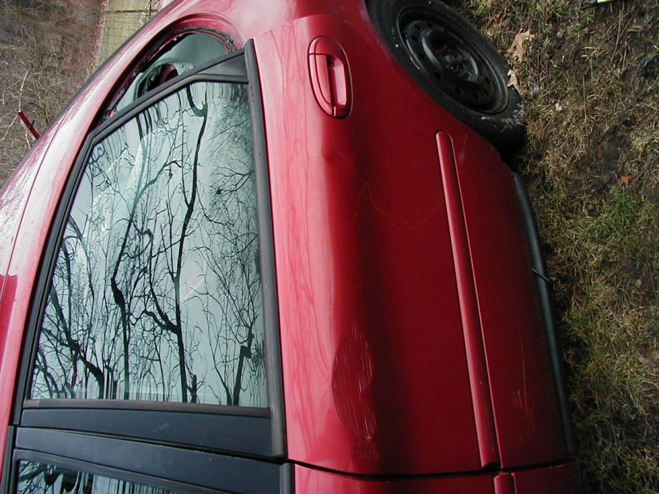 Red car door with reflections of bare trees in the window. Scratches are visible on the paint