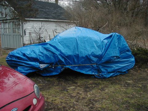 1997 Ford Taurus - A red car is parked next to a vehicle covered by a blue tarp in front of a garage.