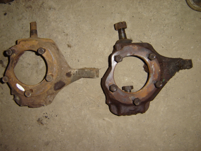 Two rusty, cast iron steering knuckles with bolts and studs on a concrete surface