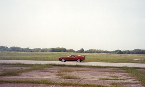 1971 Ford Mustang - A red 1971 Ford Mustang coupe is seen driving on a road with a wide open grassy field and trees in the distance under a hazy sky.
