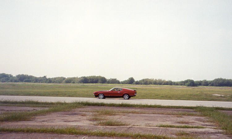 Red Mustang fastback coupe in side profile on a rural road with fields behind.