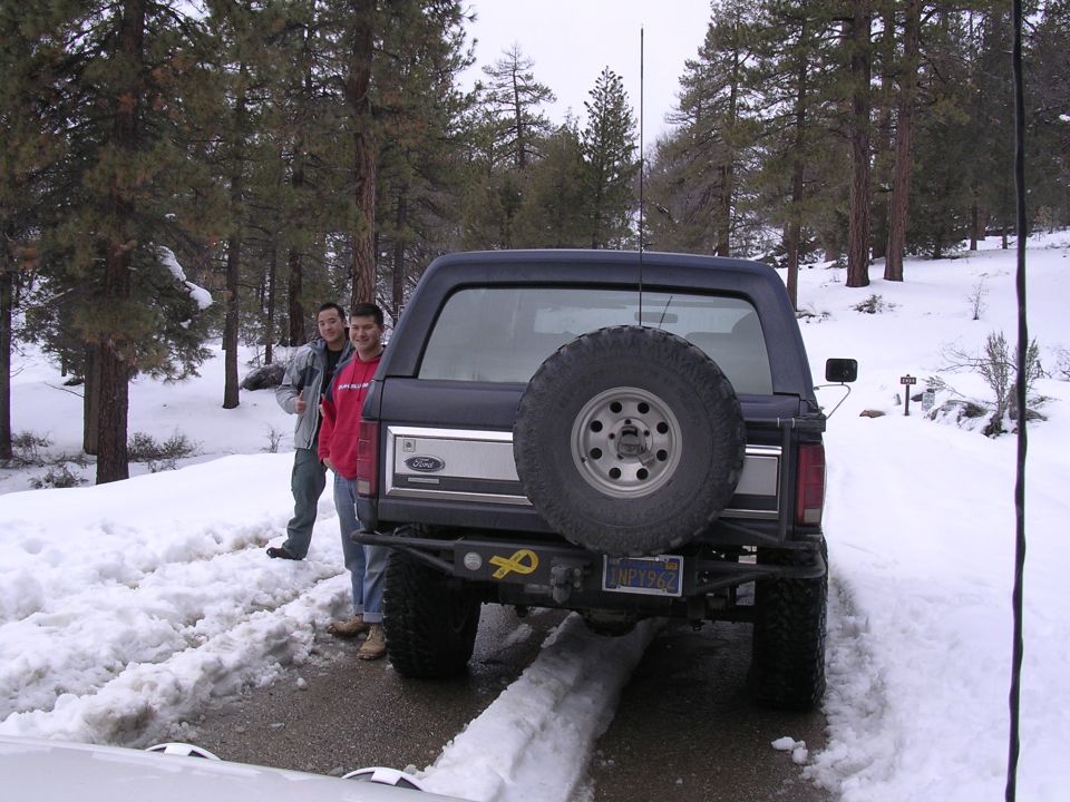 Dark blue Ford Bronco with a spare tire mounted on the rear, two men standing beside it in a snowy forest