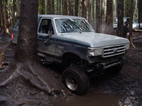 1987 Ford Bronco - This Ford Bronco is deep in the mud, surrounded by trees, with another vehicle nearby.