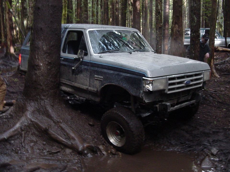Blue and silver Ford Bronco on a muddy forest trail with bumper removed and mud-terrain tires