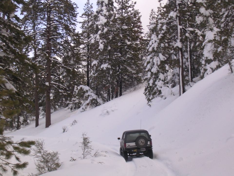 Dark Toyota pickup truck with a spare tire on the back, driving on a snowy mountain road