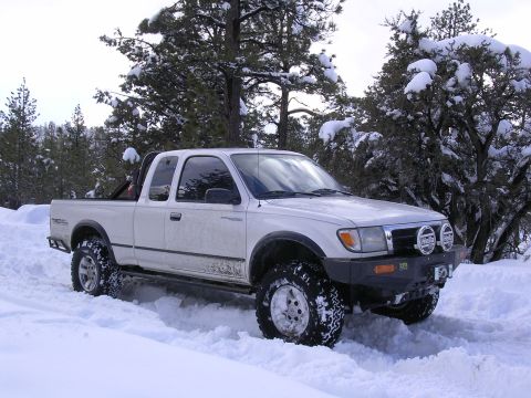 1999 Toyota Tacoma - A white 1999 Toyota Tacoma pickup truck navigates a snowy terrain, equipped with an aftermarket bumper and auxiliary lights.