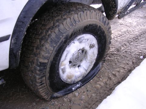 1999 Toyota Tacoma - A flat tire on a white truck is visible in snowy and muddy terrain. The hubcap shows a Toyota logo.
