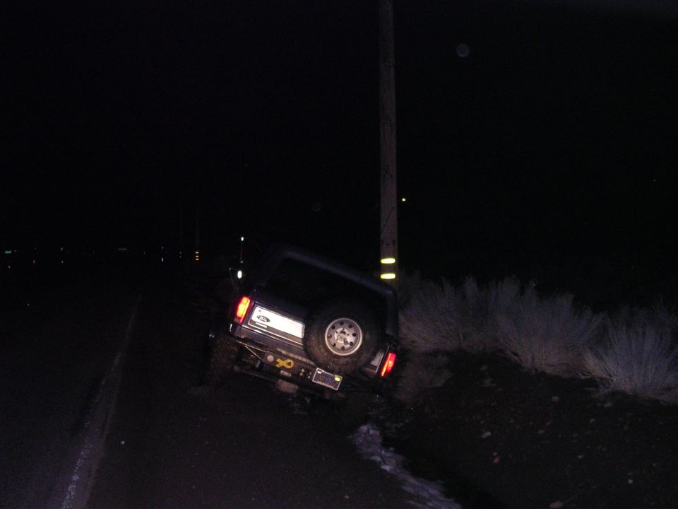 Dark SUV leaning against utility pole at night, rear view with spare tire
