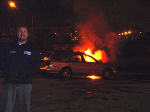 1979 Ford Bronco - A man smiles next to a silver station wagon engulfed in flames at night.