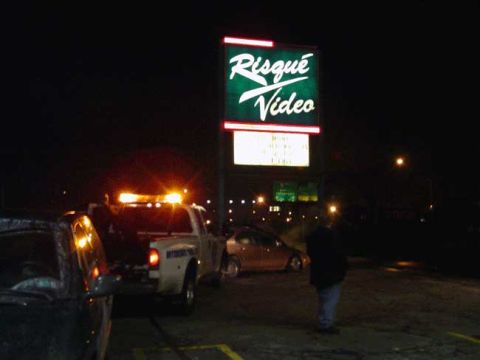 1979 Ford Bronco - Night scene in a parking lot where a white wrecker with an amber light bar hooks up a light-colored sedan beneath an illuminated "Risqué Vi…