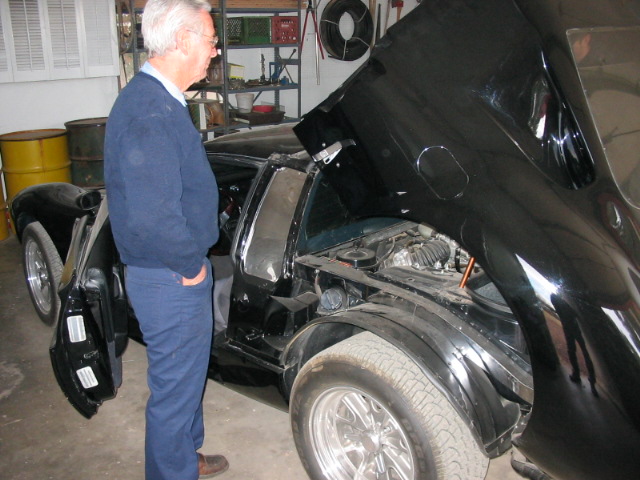 Man in navy sweater beside black sports car with front clip tilted open in a garage