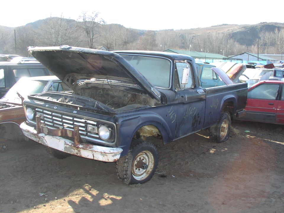 Old Ford F-series truck with hood open, parked in a junkyard. It has a flat tire and some graffiti on the side