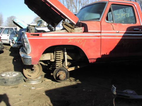 1981 Ford Bronco - A red pickup truck sits in a junkyard with its hood up and front wheels removed.