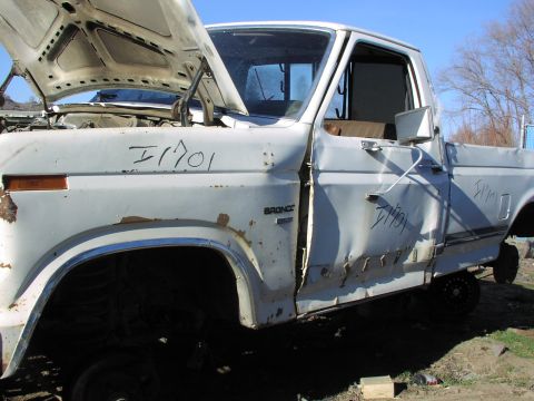 1981 Ford Bronco - This white 1981 Ford Bronco, seen in a junkyard, displays significant rust and body damage, with its hood propped open.