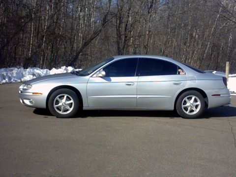 2002 Oldsmobile Aurora - Side profile of a silver 2002 Oldsmobile Aurora sedan parked on asphalt with trees and snow in the background.