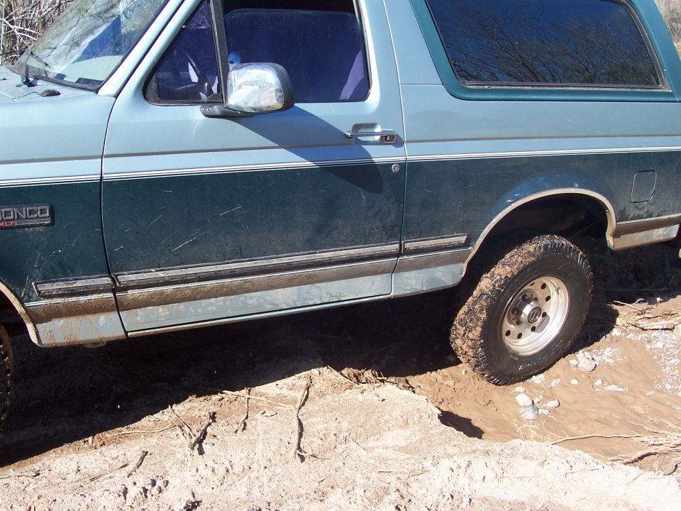 Side view of a light blue and dark green Ford Bronco stuck in mud