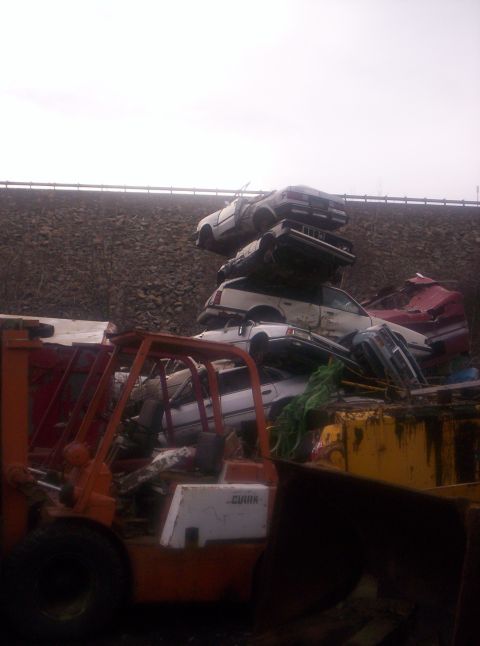 1971 Ford Mustang - A junkyard scene with multiple vehicles stacked and a forklift present.