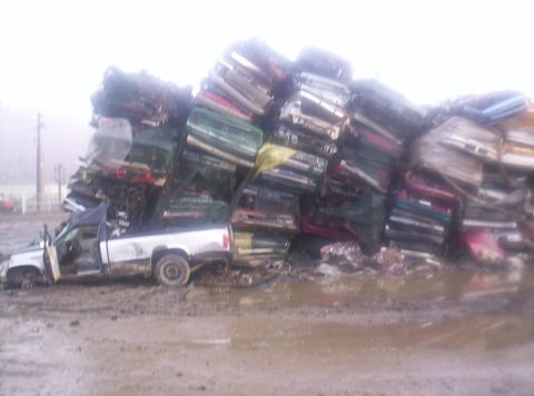 1971 Ford Mustang - A large stack of wrecked vehicles dominates the junkyard scene, with a solitary white pickup truck partially visible in the mud.