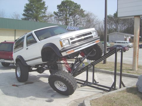 1986 Ford Bronco - White Chevrolet S-10 Blazer with extreme lift kit on a ramp, parked next to a red SUV.