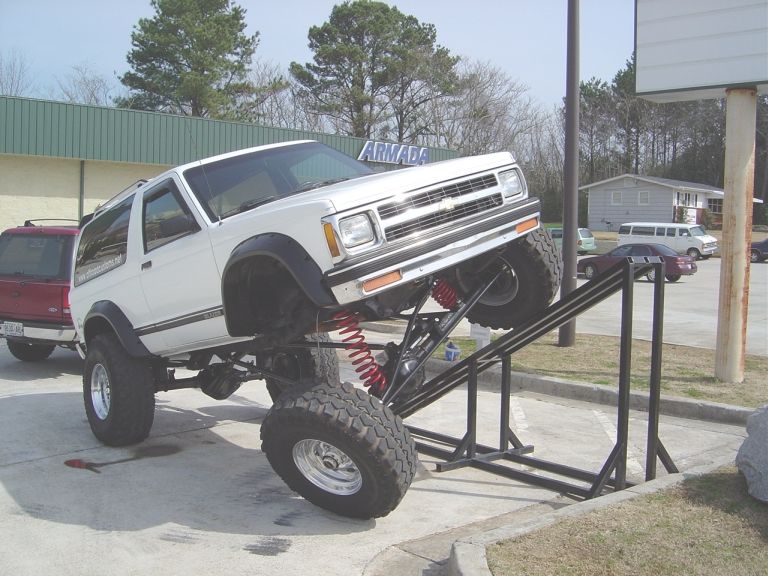 Lifted white Chevrolet S-10 Blazer on a metal ramp, red SUV in background