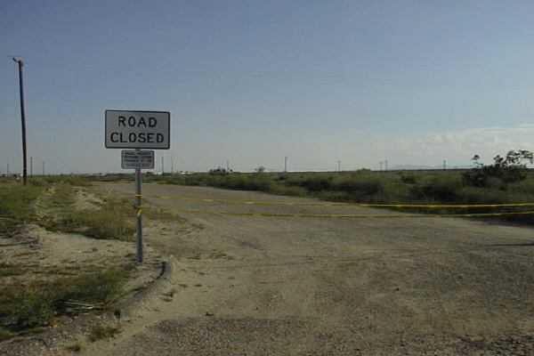 Road Closed sign and yellow caution tape across a gravel desert road