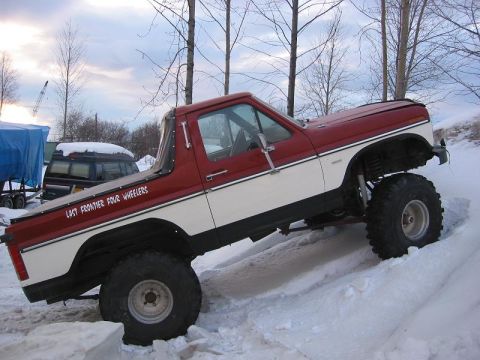 1985 Ford Bronco - A lifted red and white pickup truck navigates a snowy incline, displaying "Last Frontier Four Wheelers" on its side.
