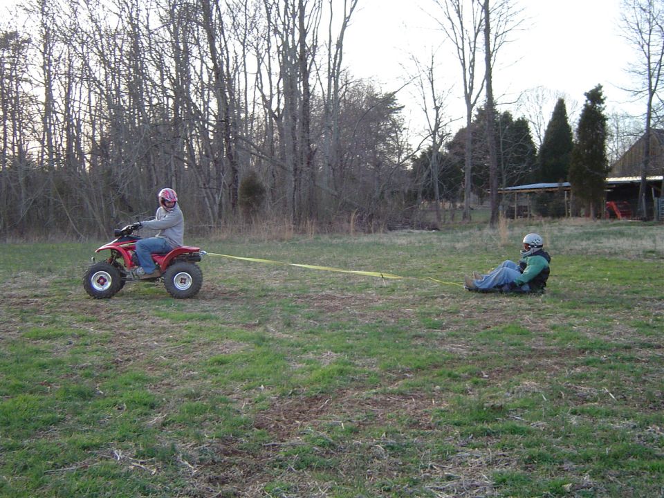 Person on red ATV pulls person lying on the ground with a yellow strap in a grassy field