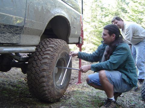 1989 Ford Bronco - A man uses a lug wrench to change a tire on a dirty, lifted SUV while another person observes.