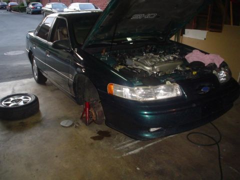 1993 Ford Taurus - A green 1993 Ford Taurus SHO is parked in a garage with its hood open and front wheel removed, indicating maintenance work.
