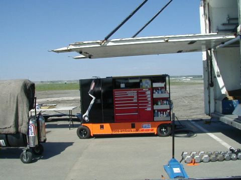 1993 Ford Probe - A mobile tool chest is parked on asphalt near a trailer and a covered cart under a bright blue sky.