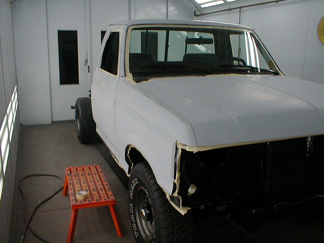 White pickup truck in spray booth with masked panels, primer prep, orange stool in foreground