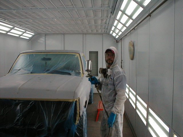 Person in white paint suit and respirator holding a spray gun beside a masked truck cab in a paint booth