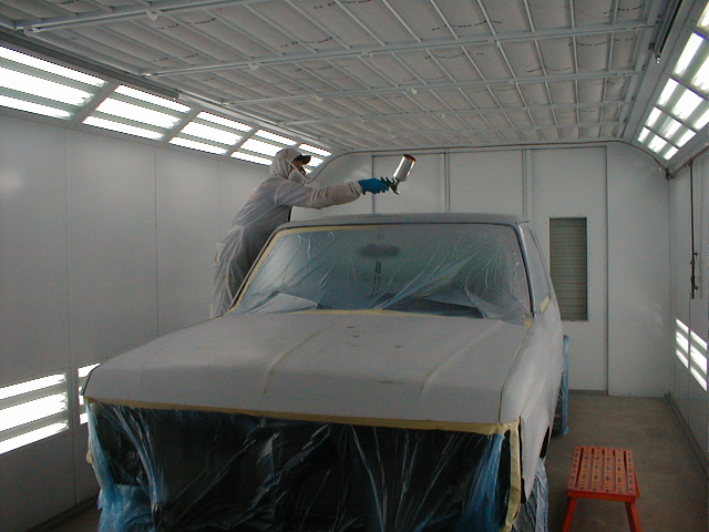 Painter in white suit reaching across the roof of a masked truck cab with a spray gun in a booth
