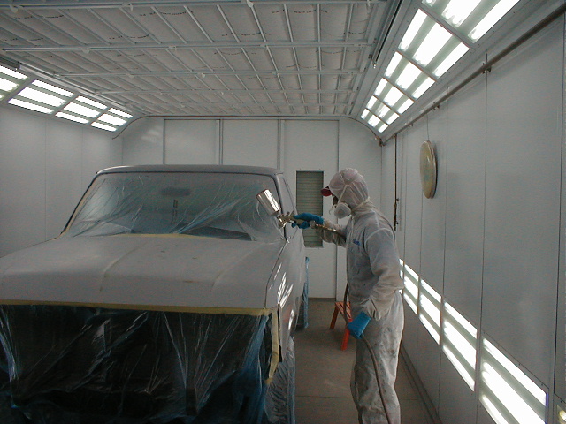 Painter in protective suit spraying a masked pickup truck in primer inside a well-lit paint booth