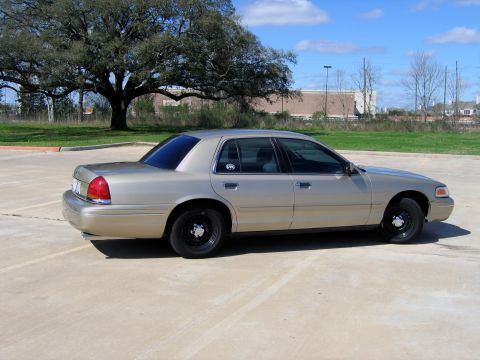 2000 Ford Crown Victoria - A gold 2000 Ford Crown Victoria is parked on a concrete lot under a large tree.