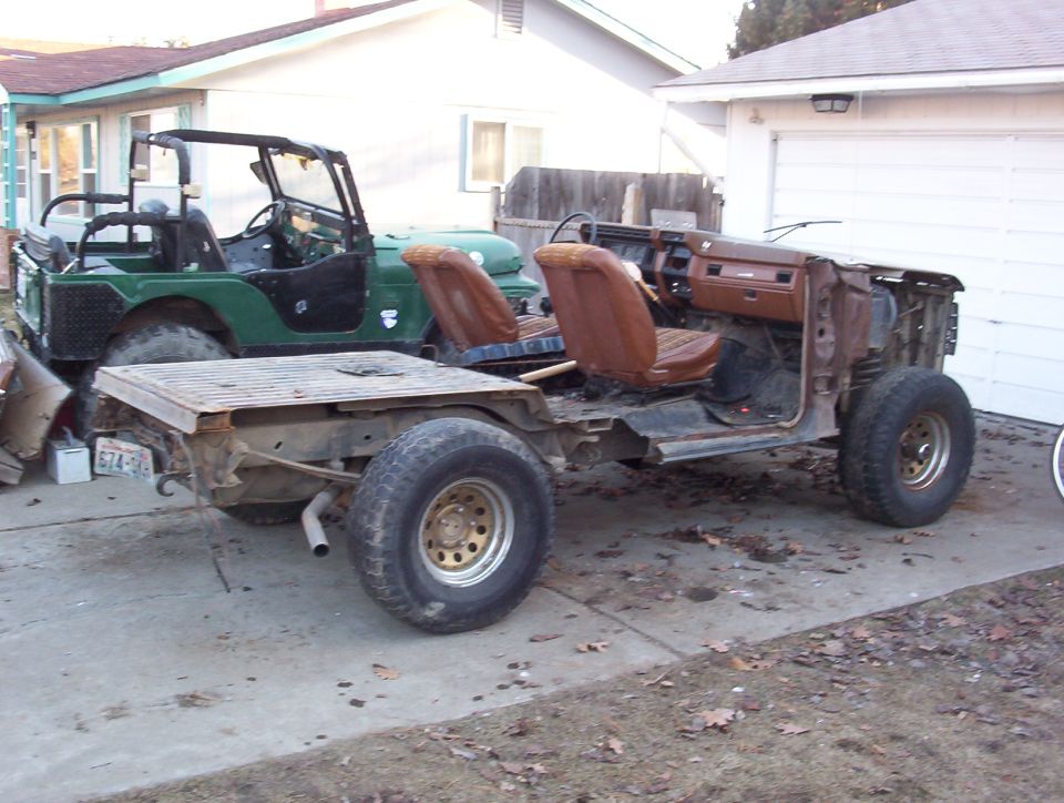 Stripped truck chassis with exposed frame and bucket seats in a driveway beside a green Jeep