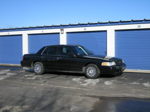 1991 Ford LTD Crown Victoria - A black sedan is parked in front of a row of blue storage unit doors under a clear sky.