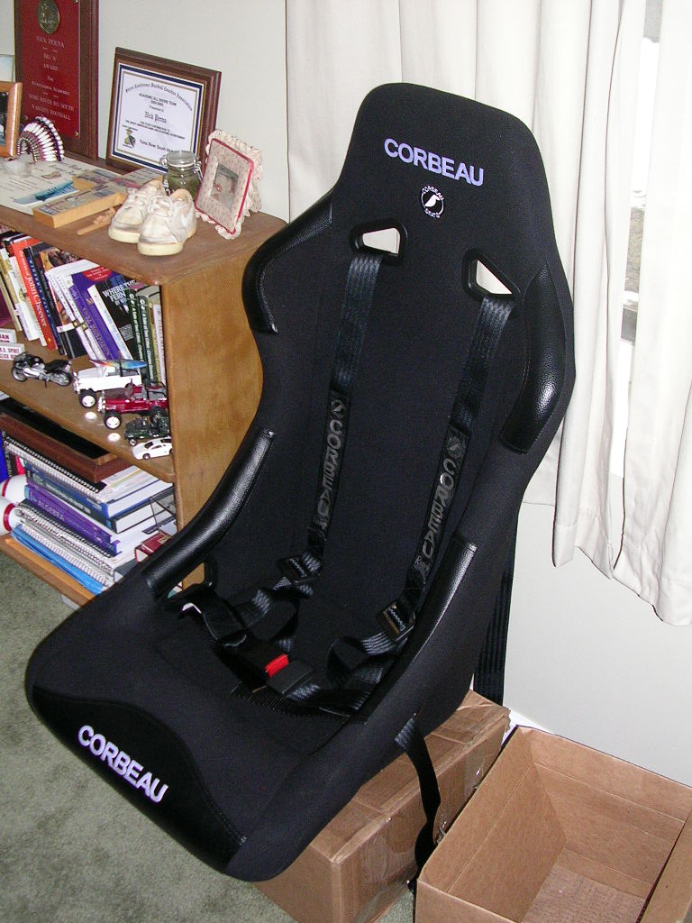 Black Corbeau racing seat with five-point harness, sitting on a cardboard box next to a bookshelf