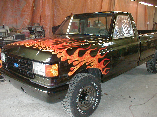 Dark green Ford F-150 truck with orange and red flame decals on the hood and fender. The truck is in a garage with orange tarps in the background