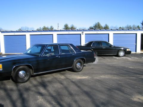 1991 Ford LTD Crown Victoria - Two sedans are parked in front of a row of blue storage unit doors under a clear sky.