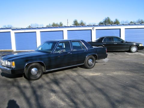 1991 Ford LTD Crown Victoria - A dark blue 1991 Ford Crown Victoria LTD sedan is parked in front of blue storage unit doors, with another black sedan behind it.