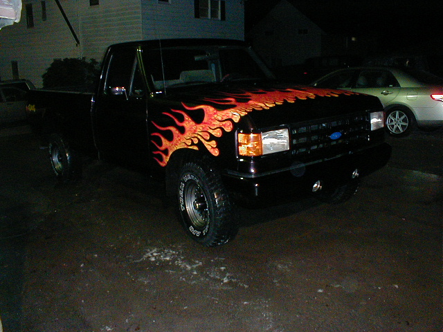 Black Ford pickup with orange flame graphics on lifted suspension and mud tires, parked at night
