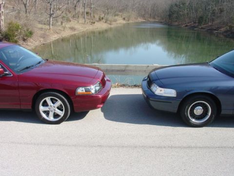1998 Ford Crown Victoria - Two sedans face each other nose-to-nose on a paved road above a green pond, with a dark blue Ford sedan on the right and a maroon/burgundy…