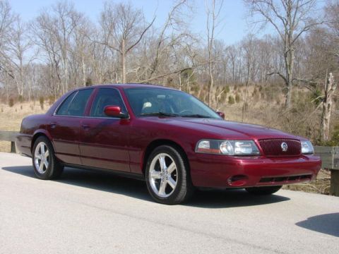1998 Ford Crown Victoria - A dark maroon/burgundy four-door sedan shown from a front three-quarter angle, parked on a paved road with bare winter trees and dry grass…