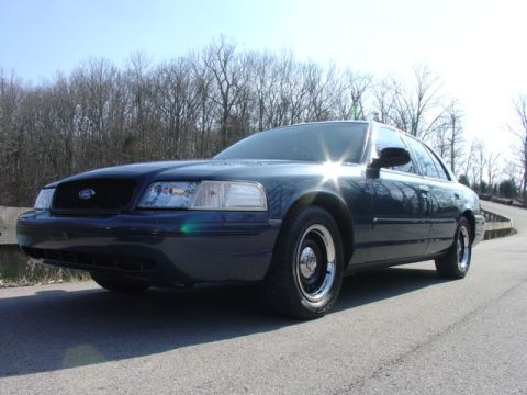 1998 Ford Crown Victoria - A dark blue/navy Ford Crown Victoria sedan photographed from a low front three-quarter angle on a paved road, with bare trees in the backgr…