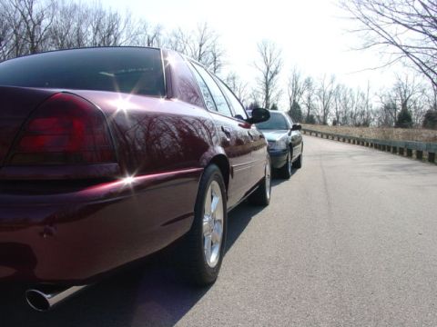 1998 Ford Crown Victoria - Low rear three-quarter close-up of a dark burgundy Ford Crown Victoria with smoked-look taillights, five-spoke alloy wheels, and a chrome e…