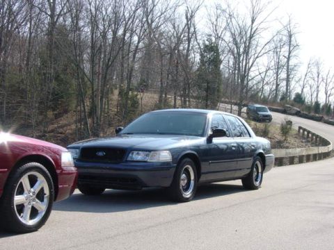 1998 Ford Crown Victoria - dark blue full-size Ford sedan travels along a rural winding road flanked by leafless trees in early spring.