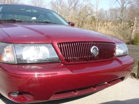 1998 Ford Crown Victoria - Close-up 3/4 front view of a maroon/dark red sedan showing the driver-side headlight and a vertical-bar chrome grille with a center emblem.…