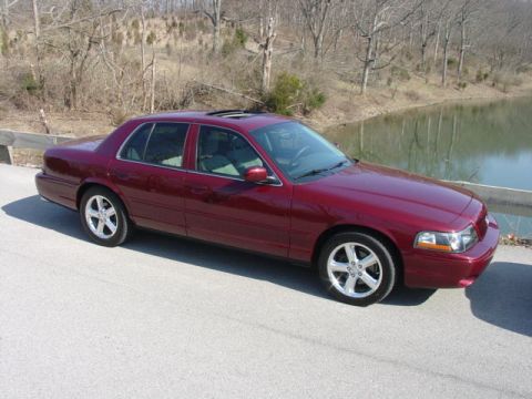 1998 Ford Crown Victoria - A dark red/maroon four-door sedan photographed from a 3/4 front angle, parked on a paved road beside a pond with bare winter trees and a gu…